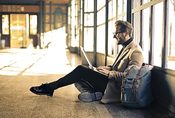 Mann trägt grauen Blazer, budapest bahnhof, Mann arbeitet Laptop, Sonnenlicht Streaming, Mann sitzt Fenster, Reiserucksack, Professionelle Kleidung, Gelegenheitsgeschäft, Innenbahn, Moderner Arbeitsbereich, Urbaner Lebensstil