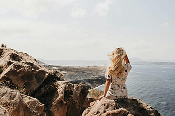 Femme assise sur une falaise, cheveux blond, falaise rocheuse, Santorin, Grèce, regardant, mer Égée, vue panoramique, photographie de voyage, beauté naturelle, aventure en plein air, moment serein