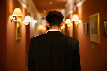 黒いスーツの男、 廊下、 暖かく照らされた、 ホテル回廊、 背面図、 歩く、 廊下の照明、 額入りの写真、 壁燭台、 屋内設定、 フォーマルな服装
