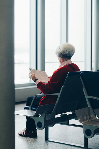 elderly woman, red coat, sitting, chair, reading newspaper, airport gate, waiting area, travel, senior citizen, indoor, airport