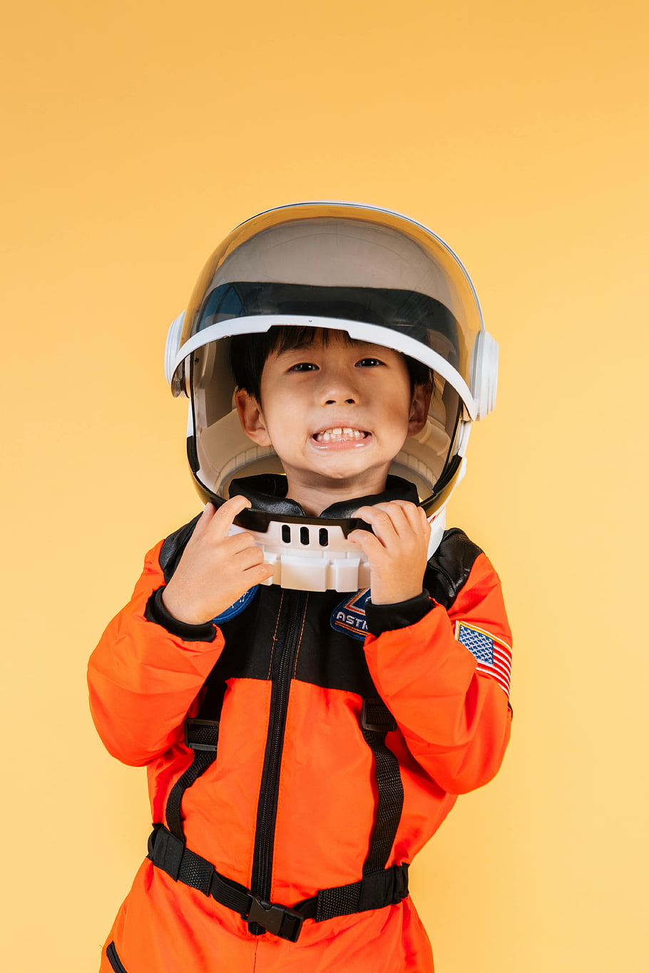 asijské chlapce, skafandr, usmívající se dítě, Astronautský kostým, Veselé dítě, žluté pozadí, kosmická helma, Oranžová skacesu, Oprava americké vlajky, průzkum vesmíru, šťastné dítě