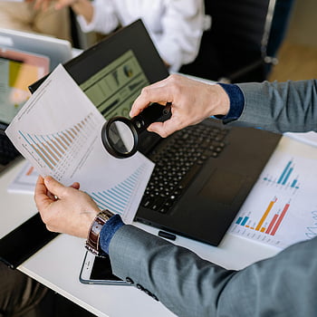 businessman, magnifying glass, data charts, laptop, office setting, examining documents, financial analysis, work environment, professional tools, business analysis, office desk