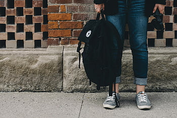 stehende Person, Tragetasche, Gelegenheitsjeans, Turnschuhe, Ziegelwand, Rucksack halten, Kamera, Outdoor-Szene, städtischen Umfeld, Freizeitkleidung, Strassenfotografie
