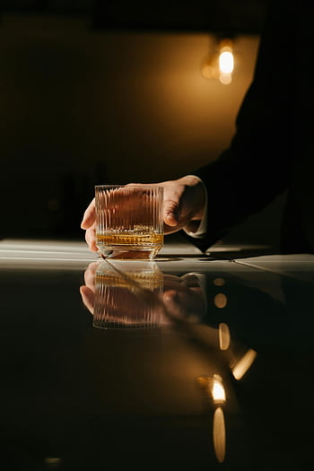 ruka drží sklo, sklenice na whisky, hnědá kapalina, slabé světlo, barový pult, odrazy, detail, čirý skleněný hrnek, osoba drží, spoře osvětlené, nastavení lišty