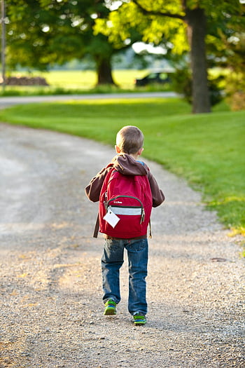 boy, brown hoodie, red backpack, walking, dirt road, tall trees, gravel path, sunny park, young boy, outdoor setting, nature walk