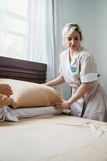 female housekeeper, fixing pillows, arranging bed, bright room, hotel room, housekeeping service, bed tidying, professional cleaner, bedroom organization, hotel staff, hospitality industry