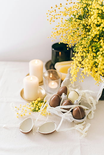 Easter-themed arrangement, chocolate eggs, candles, mimosa flowers, white table, elegant Easter, Easter decoration, spring flowers, Easter eggs, festive table setting, Easter table decor