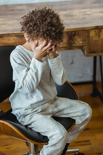 niño cubriéndose la cara, chico de pelo rizado, atuendo gris, sentado en una silla, adentro, manos cubriendo la cara, niño pequeño, pelo rizado, entorno interior, ropa casual, momento emocional
