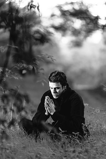 grayscale photography, man sitting, grass field, meditating, hands in prayer, black and white, outdoor meditation, peaceful scene, natural setting, contemplative pose, serene environment