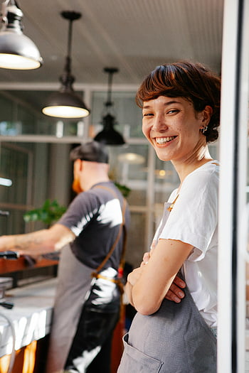 smiling barista, cheerful barista, modern cafe, welcoming atmosphere, cafeteria kitchen, unrecognizable partner, cafe setting, bright smile, modern lighting, apron, cafe worker