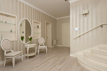 couloir classique, fauteuil, intérieur beige élégant, Papier peint à rayures, meubles élégants, atmosphère confortable, miroir rond, sol en bois, chaises blanches, escalier, étagères murales