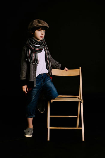 fashionable young boy, brown wooden chair, studio photography, dark background, posing with chair, stylish outfit, casual attire, studio portrait, black backdrop, wooden furniture, child fashion