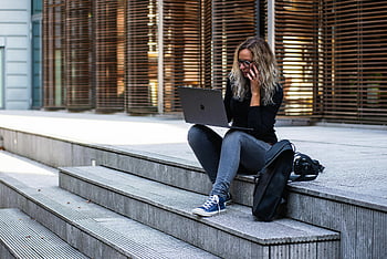 Kvinde, der sidder på trapper, bruger bærbar computer, fjernbetjening, afslappet tøj, bymæssige omgivelser, leiden, udendørs arbejde, Bærbar bruger, afslappet arbejdstøj, bymiljø, fjernbetjent