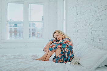 Frau, Blumenkleid, auf dem Bett sitzen, modernes Schlafzimmer, hell erleuchtet, Entspannung, weiße Mauer, großes Fenster, Freizeitpose, Innenumgebung, Bequeme Atmosphäre