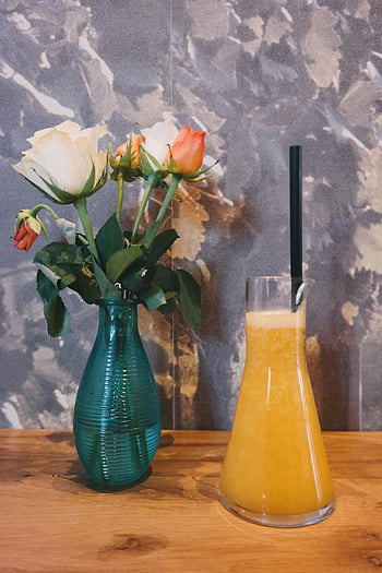 clear glass pitcher, yellow mixture, green glass vase, flowers, wooden table, orange juice, vase with roses, vibrant still life, floral arrangement, drink pitcher, table setting