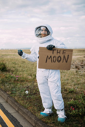 astronaut costume, hitchhiking, The Moon sign, rural road, cosplay, space suit, cardboard sign, outdoor scene, roadside, funny photo, creative photography