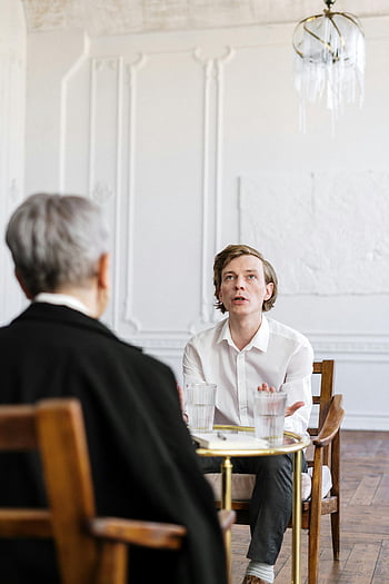 man in black suit, woman in white dress shirt, counseling session, glass table, bright loft, two people, indoor setting, professional meeting, casual conversation, modern interior, chandelier lighting