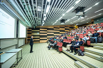 man speaking, presentation, attentive audience, modern auditorium, red chairs, speaker, presentation slide, lecture hall, professional event, conference room, audience engagement