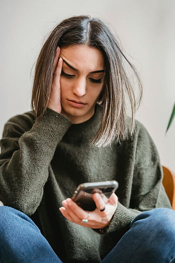 besorgte Frau, Durchsuchen Sie Smartphone, junge Frau, Smartphone lesen, Innenumgebung, besorgter Ausdruck, Legere Kleidung, Sitzposition, digitales Gerät, Mobiltelefon