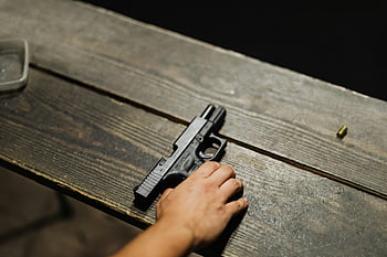 hand holding gun, black semi-automatic pistol, close-up hand, handgun, wooden table, bullet casing, firearm, pistol grip, shooting range, gun safety, firearm handling