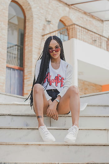 Žena sedí na schodech, módní mladá žena, městské schody, sluneční brýle, copánky, sebevědomá pozice, bílé tenisky, Neformální outfit, panoráma pozadí, moderní architektura, pouliční styl