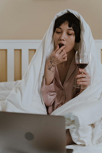 Asian woman, pajamas, wine glass, chocolate, sitting in bed, wrapped in blanket, enjoying wine, cozy moment, relaxation, laptop, comfort food