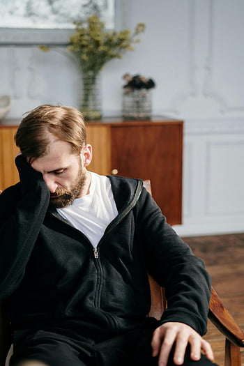 man, black zip up jacket, sitting, chair, bearded, hoodie, indoors, deep in thought, casual attire, contemplative, home setting