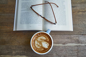 white ceramic mug, eyeglasses, open book, wooden table, cappuccino, warm scene, coffee cup, reading glasses, cozy setting, tabletop arrangement, latte art