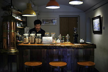 Café, Barista, warmes Interieur, Atmosphäre einladende, Getränkevorbereitung, Bartheke, Schwarzes Poloshirt, Holzhocker, Coffee-Shop-Interieur, Barista hinter der Theke, gemütliches Ambiente