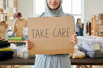 gray hijab, brown cardboard, take care sign, smiling volunteer, charity event, indoors, volunteer work, community service, donation center, humanitarian aid, charity sign
