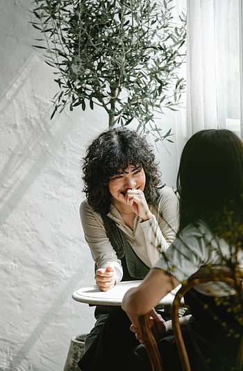 Positive ethnische Kollegeninnen, Tabelle im Café, Zwei Frauen lachen, Kaffee, Sunny Café -Einstellung, weibliche Freunde, Gelegenheitsgespräch, Innencafé, natürliches Licht, Entspannte Atmosphäre, soziale Interaktion