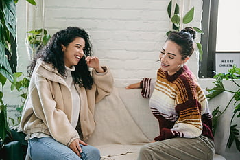 Femei tinere fericite, așezat pe canapea, vorbind, Setare interioară confortabilă, Verderie luxuriantă, Conversație caldă, doi prieteni, zâmbitor, haine casual, decoratiune interioara, plante