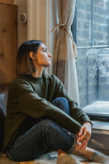 donna sognante, gambe incrociate, seduto vicino alla finestra, giovane donna, maglione, Guarda pensieroso, abbracciare la solitudine, Scena indoor, Abbigliamento casual, Posa rilassata, umore contemplativo