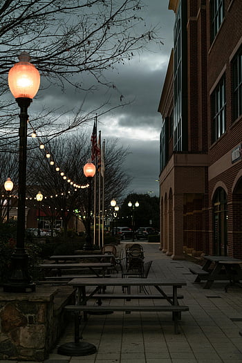 street lamps, string of lights, urban evening, moody scene, deserted street, glowing lights, building exterior, outdoor seating, picnic tables, evening atmosphere, cityscape
