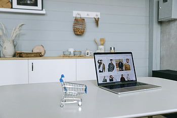 Laptop, Online Einkaufen, Miniatureinkaufswagen, minimaler Schreibtisch, Einkaufsportal, E-Commerce, Warenpräsentation, digitaler Einzelhandel, Innenministerium, Moderner Arbeitsbereich, Online-Shop