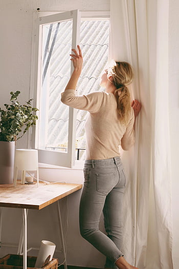 Frau schaut aus dem Fenster, Sonnenlicht, gemütliches Zimmer, minimalistischer Raum, Fenster öffnen, Innenszene, natürliches Licht, Lässige Kleidung, Wohnkultur, Schreibtischlampe, Pflanze