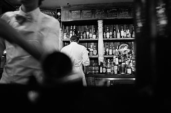 grayscale photography, man standing, cabinet, bartenders at work, vintage bar setting, black and white photo, bar scene, vintage bar, bartender, bar cabinet, alcohol bottles
