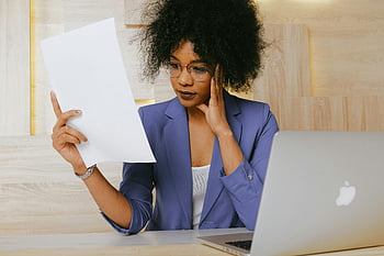 woman in blue blazer, holding white paper, businesswoman, working on laptop, reviewing document, office setting, professional attire, desk work, focused expression, document review, office environment