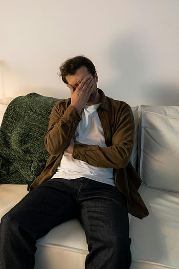zmartwiony człowiek, siedząc na kanapie, oznaki niepokoju, niepokój, w domu, stres emocjonalny, zdrowie psychiczne, sofa, ustawienie wewnętrzne, codzienne ubrania, biała kanapa