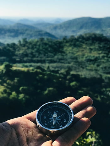 handheld compass, round compass, grey and black compass, majestic green mountains, blue skies, travel themes, outdoor navigation, nature backdrop, adventure photography, scenic landscape, travel compass